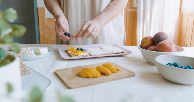 Woman making sweet sandwiches with crispbread, cream cheese, blueberry, slices of peach, coconut flakes and honey for summer picnic or brunch.