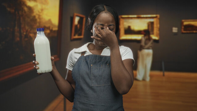 Woman wearing apron holds milk bottle and pinches nose, eyes closed, in an art gallery building with framed paintings; disgust reaction.