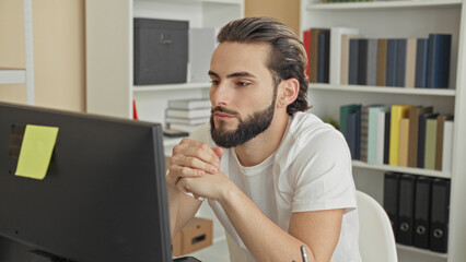 Fototapeta premium Man young hispanic at computer with hands covering face and rubbing jaw while seated at desk in building; stress concentration.