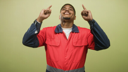 Fototapeta premium Man in red work uniform pointing both index fingers upward, smiling with beard visible, wearing coveralls under studio lighting; celebration achievement.