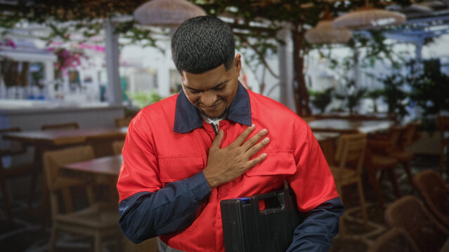 Man worker in red uniform holding toolbox places hand on chest inside building dining area with tables chairs plants and hanging lamps; pride duty.