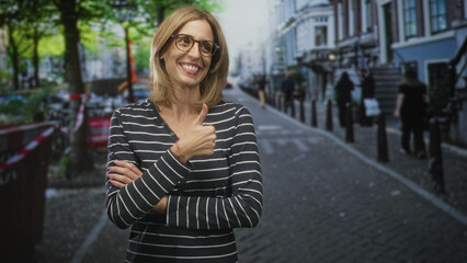 Fototapeta premium Woman with crossed arms showing hands and eyeglasses standing on a cobbled street near bollards and trees; confidence observation reflection.