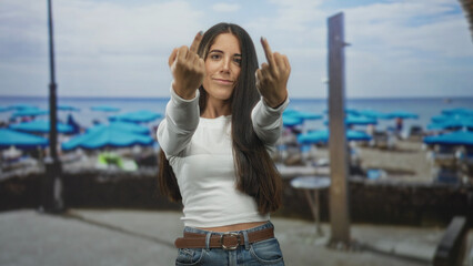 Obraz premium Woman raising middle finger with bare hand on a street by crowded beach umbrellas and ocean shore; defiance.