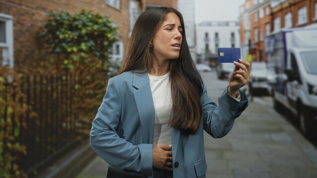 Young woman holds a blue creditcard in her hand while clutching her stomach and frowning on a city street lined with vans and brick buildings; financial frustration.