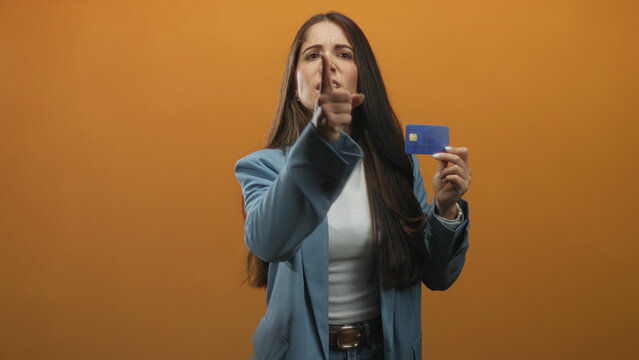 Woman holding blue creditcard and raising fist toward camera in studio orange backdrop while pointing with other hand; frustration consumer rights.