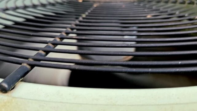 A black air conditioning fan spinning outdoors in summer on an older unit, showing the protective safety grille and condenser housing. Video
