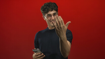 Man holding a smartphone and glancing down in a bright red studio while wearing glasses and a black shirt  curiosity engagement. © Krakenimages.com