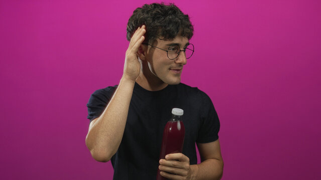 Man wearing glasses and a dark t shirt holding a bottle of juice, hand to ear and smiling in studio; taste test playful.