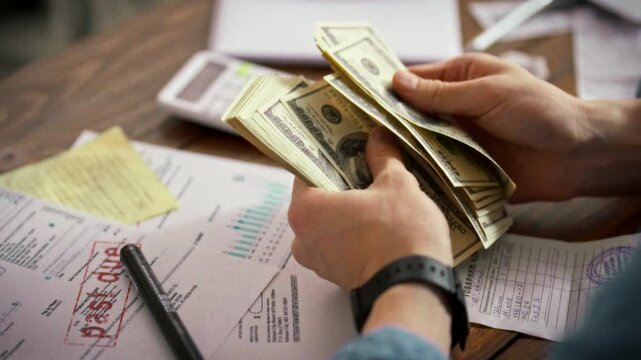 A young businessman's hands tally banknotes beside unpaid bills and scattered documents, the weight of finance pressing down. The scene conveys mounting pressure as he confronts growing obligations