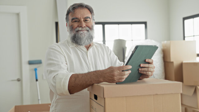 Man tapping tablet on a cardboard box while organizing packed moving packages inside a bright building with stacked boxes and bubblewrap; moving planning serenity.