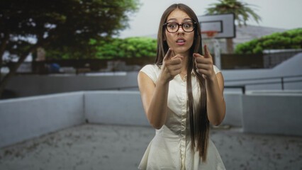 Fototapeta premium Woman with long hair and glasses pointing fingers toward camera by a basketball hoop in a building courtyard; surprise curiosity invitation.