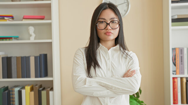 Woman wearing glasses with arms crossed and unsmiling gaze in a home office lined with books and a clock; confidence determination.