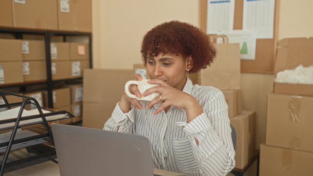 Woman holds coffee mug with both hands, eyes closed and smiling at laptop, surrounded by packed cardboard boxes, shipping labels and parcel shelves in building; small business calm.