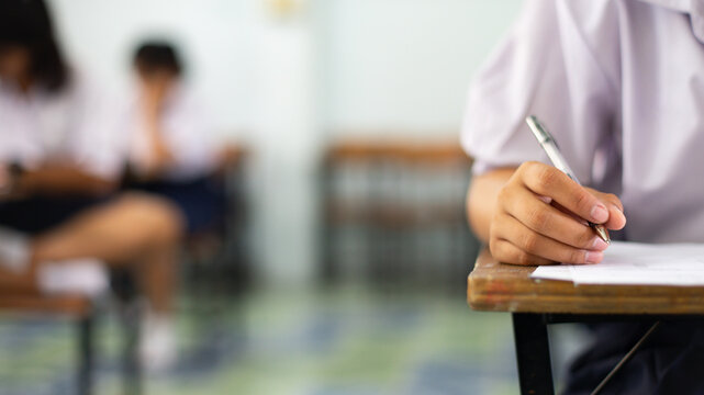 Close-up of a student's hand diligently writing on an exam paper during a school test. 
The student is using a professional pen , sitting at a wooden desk in a brightly lit classroom.