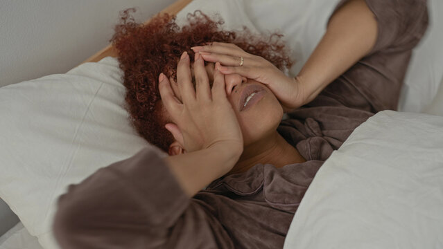 Woman covering face with both hands while lying on bed in studio setting, curly hair visible and expression strained; fatigue.