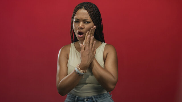 Young african american woman with hand on cheek for toothache in studio against red backdrop, jaw clenched and open mouth; pain discomfort.