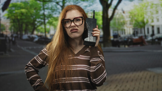 Redhead woman holding vintage cellphone to ear, raising hand on street, wearing striped brown sweater and glasses; retro curiosity confusion.