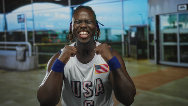 Young man smiling with clenched fists, wearing usa basketball jersey with flag patch and blue wristbands, dreadlocks visible, standing at airport terminal entrance; pride celebration.
