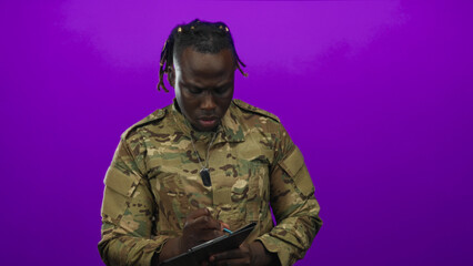 Fototapeta premium Young man in camouflage military uniform with dog tags writes on tablet with stylus and visible hands in purple studio; determination.