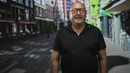 Senior hispanic man smiling, wearing glasses and black polo shirt, stands facing camera on a street lined with shops and cafes  friendly contentment. © Krakenimages.com