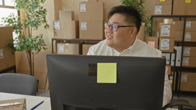 Man at desktop computer smiles while looking to right among stacked parcels in packing building; small business productivity contentment.