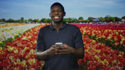 Fototapeta premium Man holding smartphone with both hands, smiling and typing while standing before tulip field in studio set; joy connection.
