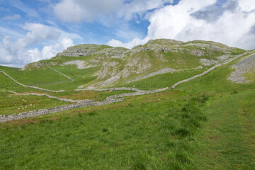Limestone hills of Warrendale Knotts rise above lush green pastures partitioned by traditional dry stone walls under a cloudy sky in Settle, Yorkshire Dales National Park, England, UK. © Matthew