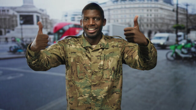 Man soldier in camouflage giving thumbs up, hands raised and smiling on a busy urban street with vehicles and a monument visible; pride duty.