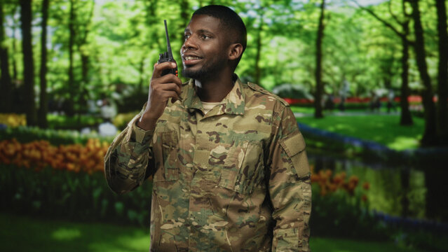 Man soldier speaks into radio in a forest wearing camouflage uniform, holding a handheld transceiver and scanning the area with a slight smile; duty vigilance.