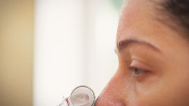 In bathroom, girl wrapped in a white robe with a towel on her head uses a derma roller for microneedling, staring at the mirror as the skincare tool stimulates collagen and brightens her facial tone