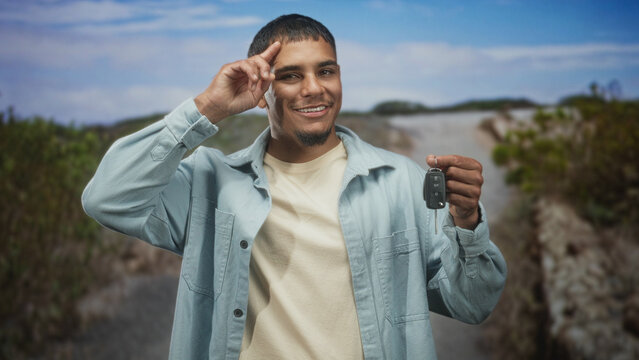 Man holding car keys with finger to temple salute on a street by dunes, smiling broadly; confidence success.