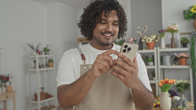 Man florist tapping smartphone with hands in a flower shop building, wearing apron and smiling at screen; small business care cheerful.