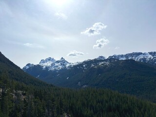 Fototapeta premium Snow Capped Mountain Peaks Overlooking Dense Pine Forest Valley, Beautiful Alpine Wilderness Landscape, Sunny Day Hiking Travel Background in Canada
