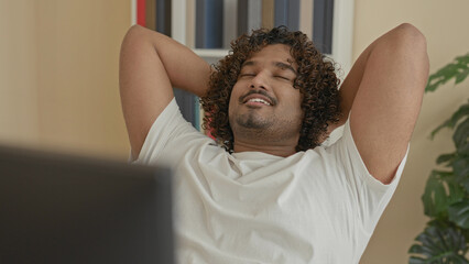 Young indian man at computer, hands behind head and leaning back smiling in a building office with bookshelves and plant visible  relaxation. © Krakenimages.com