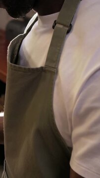 Vertical video: Camera pulling back showing male in olive apron leaning on counter, prepping food