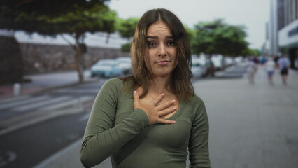 Fototapeta premium Young woman with hand on chest wearing green shirt and necklace on city street pedestrian walkway; regret apology.