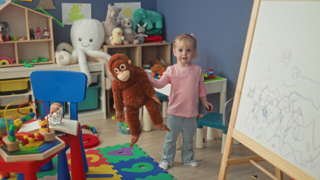 Little blonde girl hugging a large stuffed monkey with hands in a studio playroom; playful innocence.