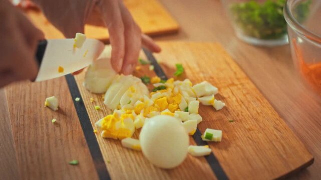 Cutting eggs on wooden cutting board, a young housewife prepares a meal in her kitchen. She skillfully uses a sharp knife, handling the eggs with care, as she works through her daytime cooking tasks