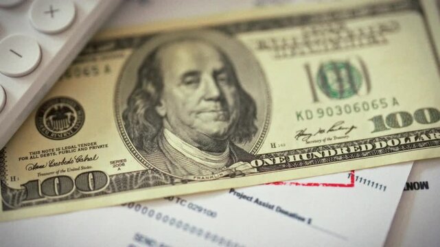 On a cluttered office desk, a close-up shows a hundred-dollar bill beside a calculator on a stack of office papers, highlighting precise money management and finance planning in everyday budgeting