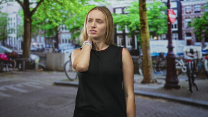 Obraz premium Woman touching neck in a black tanktop on a city street with bicycles and lampposts visible; quiet contemplation.