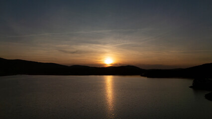 Obraz premium Tranquil Dusk Over Water: The sun dips below the horizon, casting a golden reflection on the calm water surface, with silhouetted hills framing the serene twilight scene.