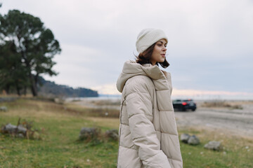 Woman in a beige puffer coat and knit beanie stands outdoors by a coastal field under an overcast sky, with distant water and a blurred road © SHOTPRIME STUDIO