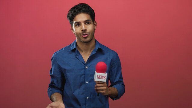 Man holding microphone with red windscreen and gesturing open palm while speaking in studio with pink backdrop; informative calm.