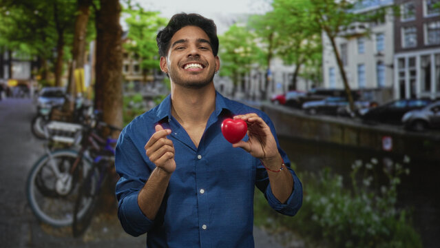 Young man in a blue shirt holding a red heart and making a finger heart gesture on a street, smiling; romance connection.