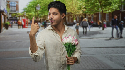 Obraz premium Young hispanic man smiling, holding pink tulip bouquet with hands in front, standing on a city street with blurred pedestrians; romance connection.