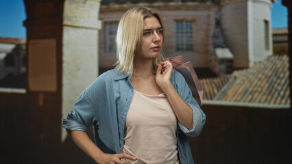 Fototapeta premium Woman holding pink shopping bag on shoulder with hand on hip in studio set with a projected building backdrop; pensive reflection.