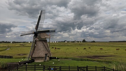 Aerial view of the traditional Borgmolen windmill in Earnewald, surrounded by vibrant green meadows and wetlands under a dramatic cloudy sky, with distant sailboats. © Jens