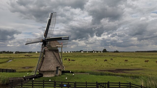 Aerial view of the traditional Borgmolen windmill in Earnewald, surrounded by vibrant green meadows and wetlands under a dramatic cloudy sky, with distant sailboats.