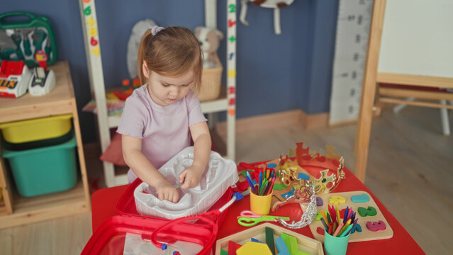 Toddler girl sorting toy pieces with hands on a red play table inside a preschool building; curiosity imaginative play.