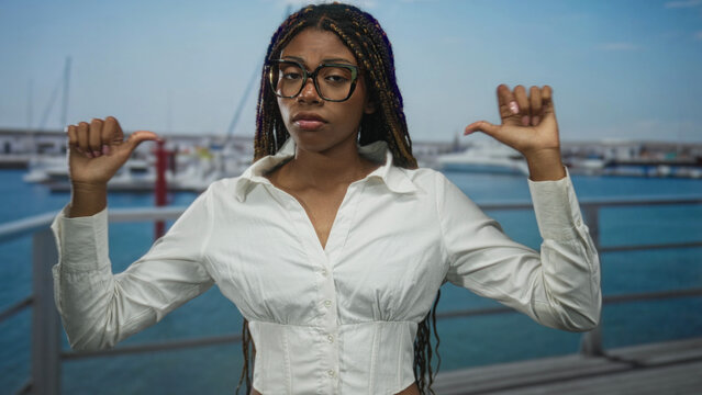 Woman points thumbs to chest at port boardwalk wearing white blouse and glasses, braids visible; confidence.